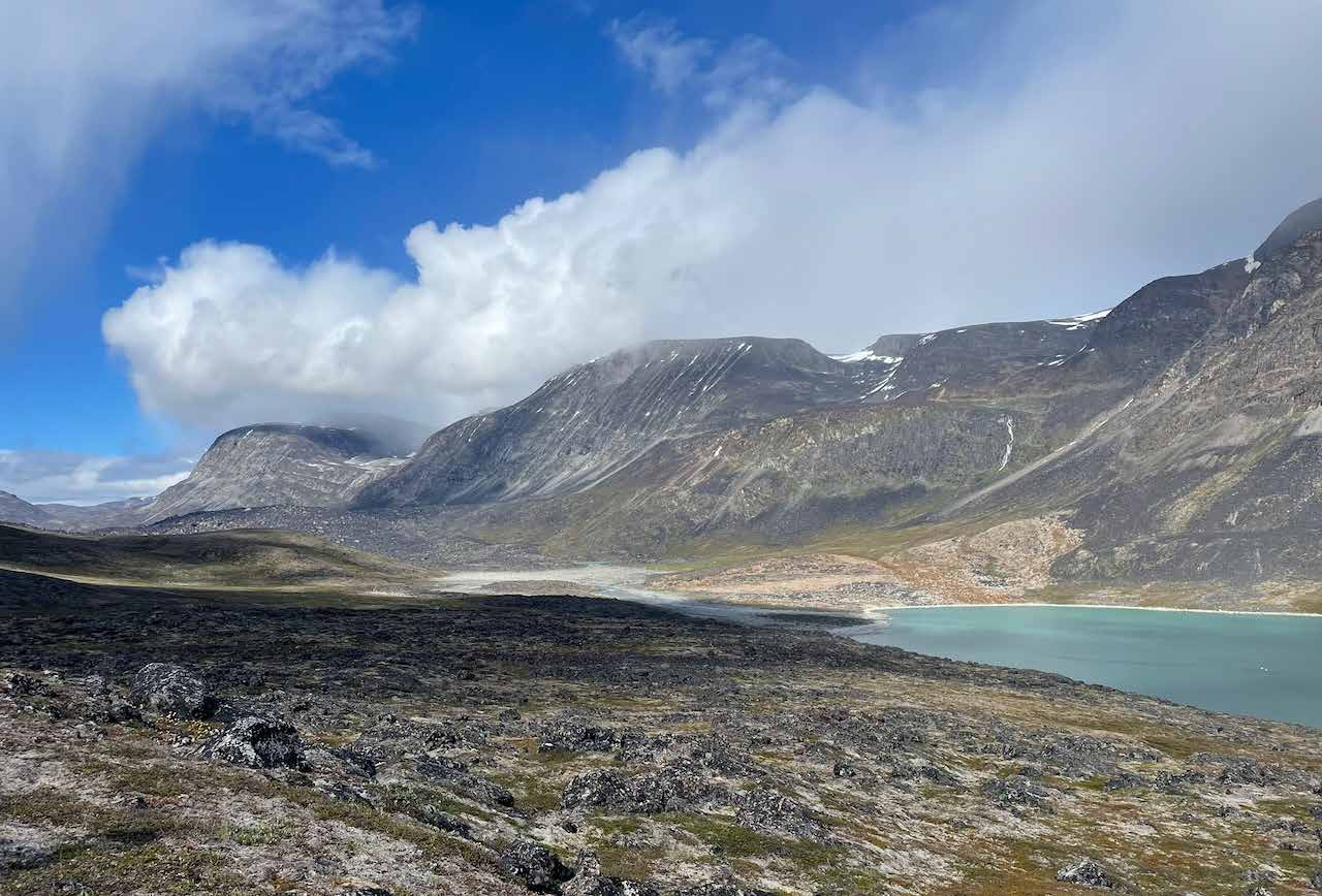 Weite Tundralandschaft mit dunklem Gestein, dahinter Berge und eine türkisfarbene Bucht