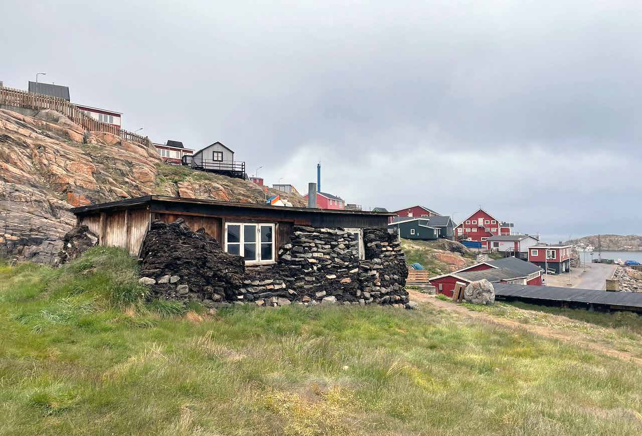 Historisches Haus aus Torfsoden und Steinen in Uummannaq, dahinter der Ort und der Hafen